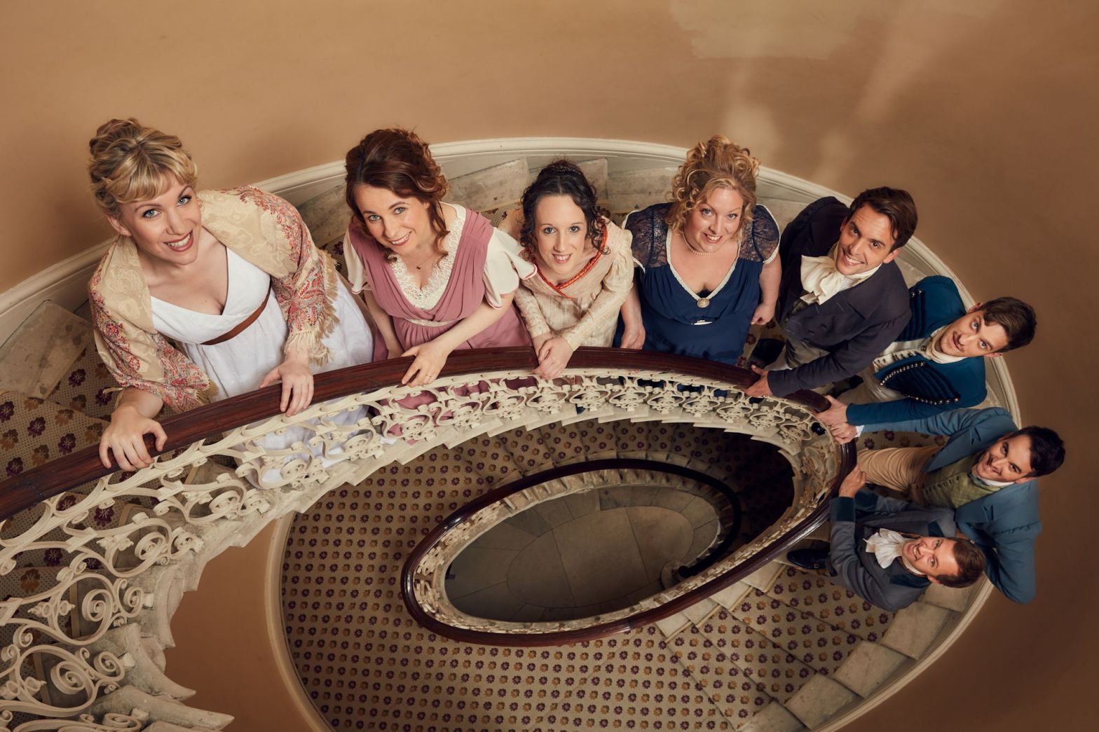 Eight performers in regency costumes line up on a spiral staircase.